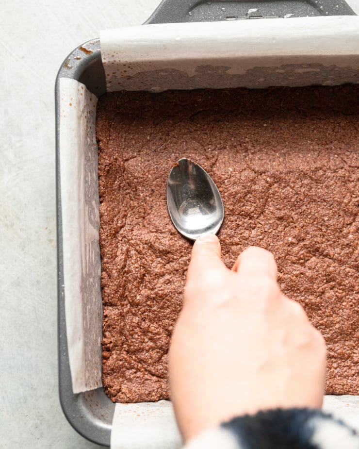 Top view showing chocolate bark baking in a square pan. Use the back of a spoon with one hand to press down on the crust and allow it to harden slightly.
