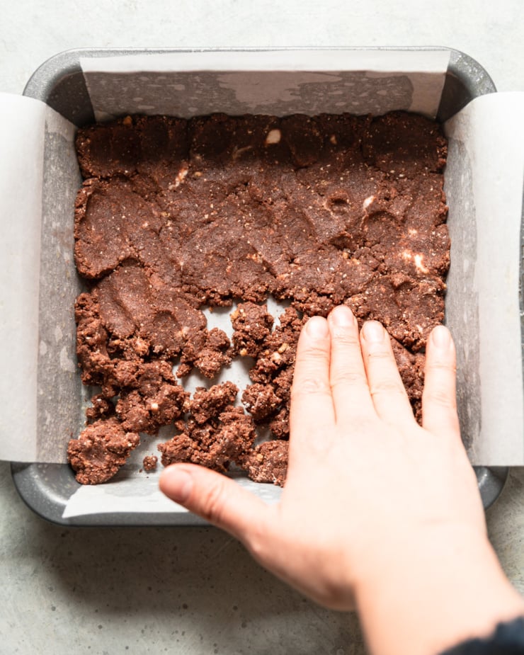 An overhead shot shows a hand pressing the chocolate bark mixture into a pan lined with parchment paper.