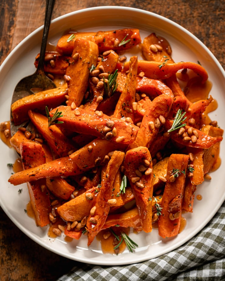 An overhead shot shows roasted honeynut squash wedges on a plate, topped with toasted pine nuts, herbs, and apple cider reduction. A check linen napkin is nearby and a silver serving spatula is sticking out of the squash.