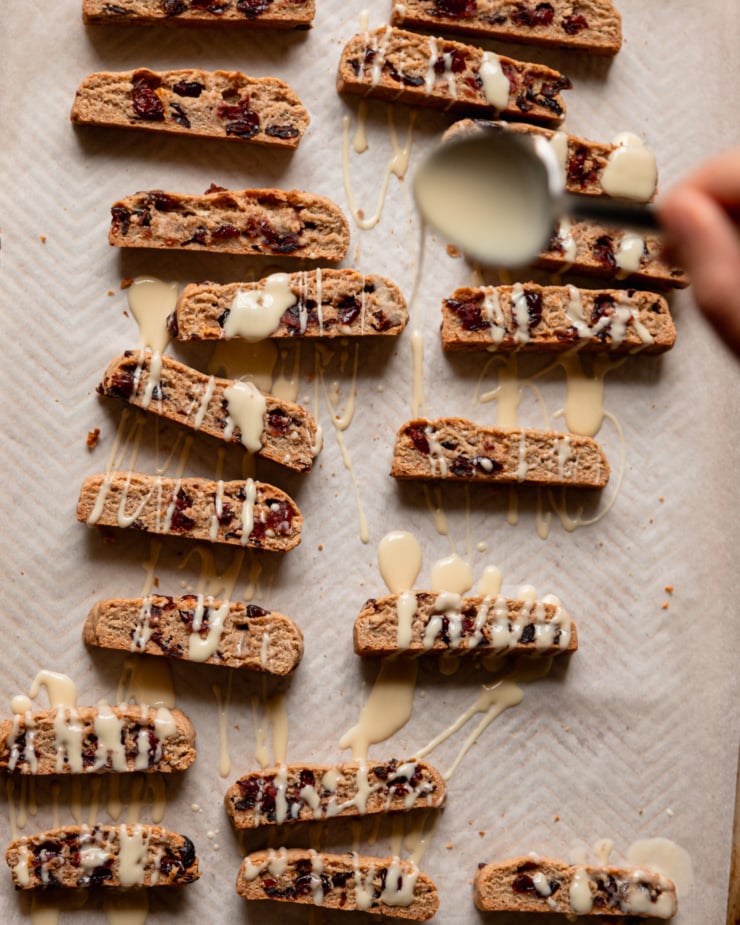 An overhead photo shows a hand using a spoon to drizzle white chocolate over a vegan orange-cranberry shortcake.