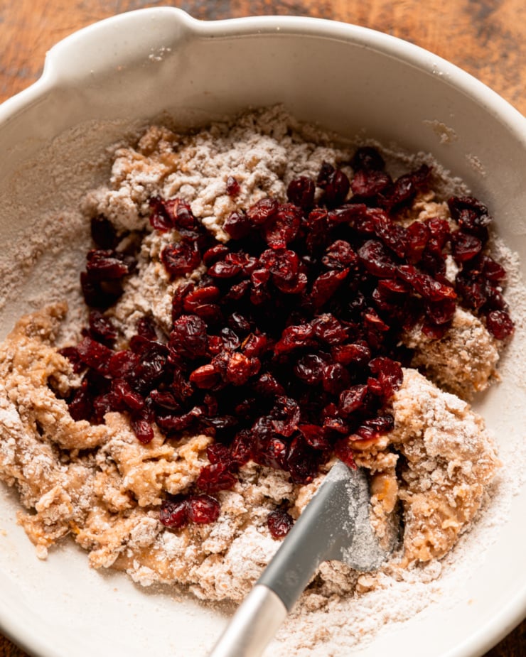 An overhead view shows cookie batter in a bowl with a pile of dried cranberries on top.