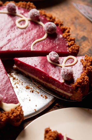 A 3/4 angle image shows a slice being taken out of a vegan cranberry tart. You can see a defined white chocolate bottom layer with the cranberry curd layer on top. The top is garnished with melted white chocolate and sugared cranberries.