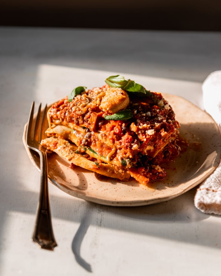 The 3/4 angle shot shows a piece of vegan lasagna on a plate with a fork next to it. This photo was taken in direct sunlight.