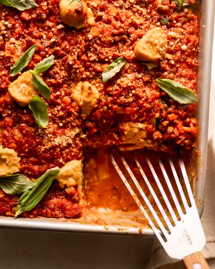 A close-up overhead shot shows vegan lasagna baking in a baking dish. Top with a dollop of tofu cashew ricotta, fresh basil leaves and veggie 