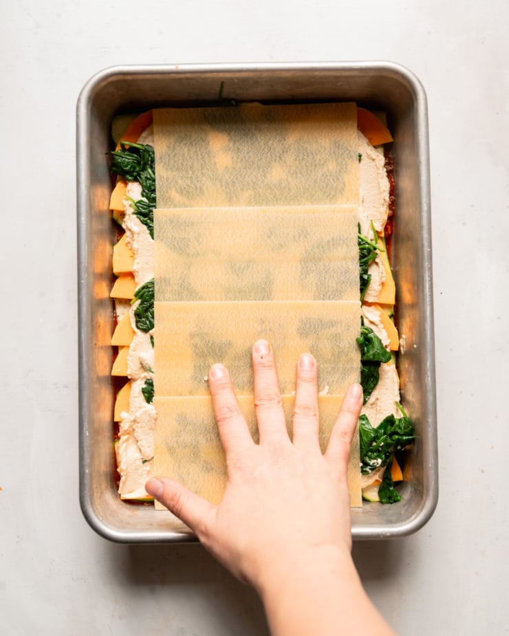The top-down photo shows a hand pressing lasagna noodles into a baking pan filled with sliced ​​vegetables and tofu-cashew ricotta ganache.