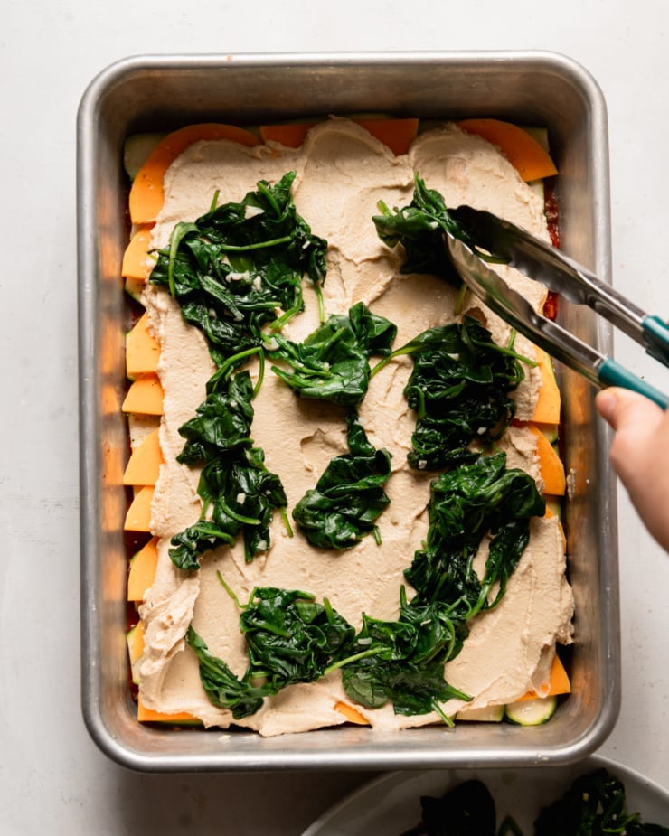 The overhead photo shows a hand using tongs to hold sautéed baby spinach atop a mixture of tofu, cashew ricotta and sliced ​​vegetables in a baking dish.