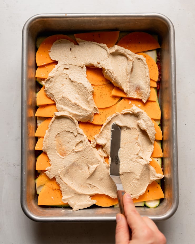 Top-down photo shows a hand using an offset spatula to spread tofu cashew ricotta ganache over some thinly sliced ​​vegetables in a baking dish.