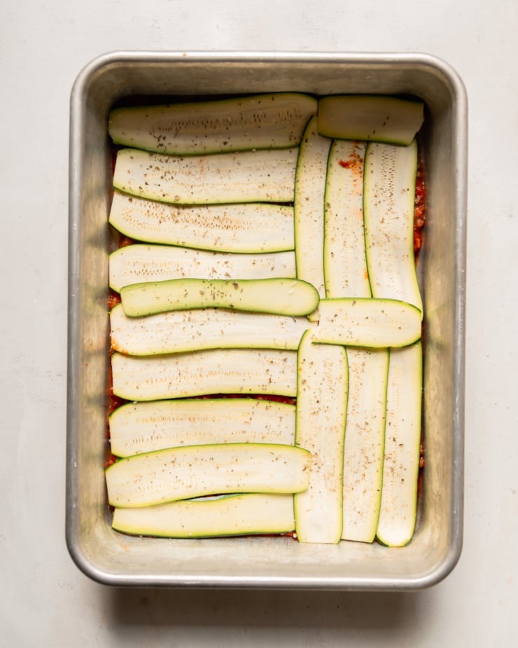 An overhead photo shows thinly sliced ​​zucchini in a baking dish evenly covered with a layer of tomato sauce.
