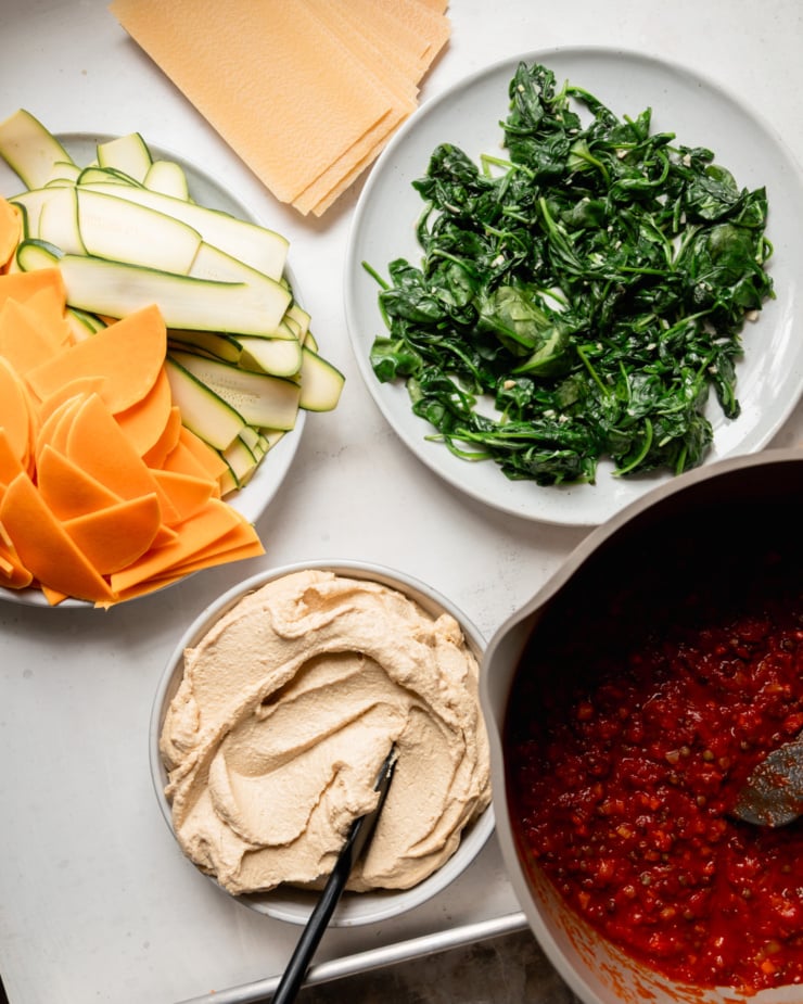 An overhead view shows the components ready to make vegan lasagna: lasagna noodles, sautéed spinach, lentil and vegetable tomato sauce, tofu cashew ricotta, and thinly sliced ​​butternut squash and zucchini.