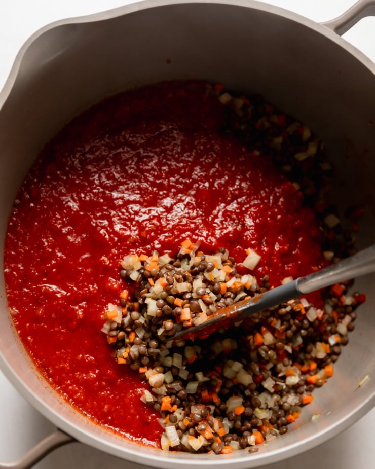 Top view showing a pot of sautéed diced vegetables, cooked lentils and tomato paste. A spatula is stirring everything up.