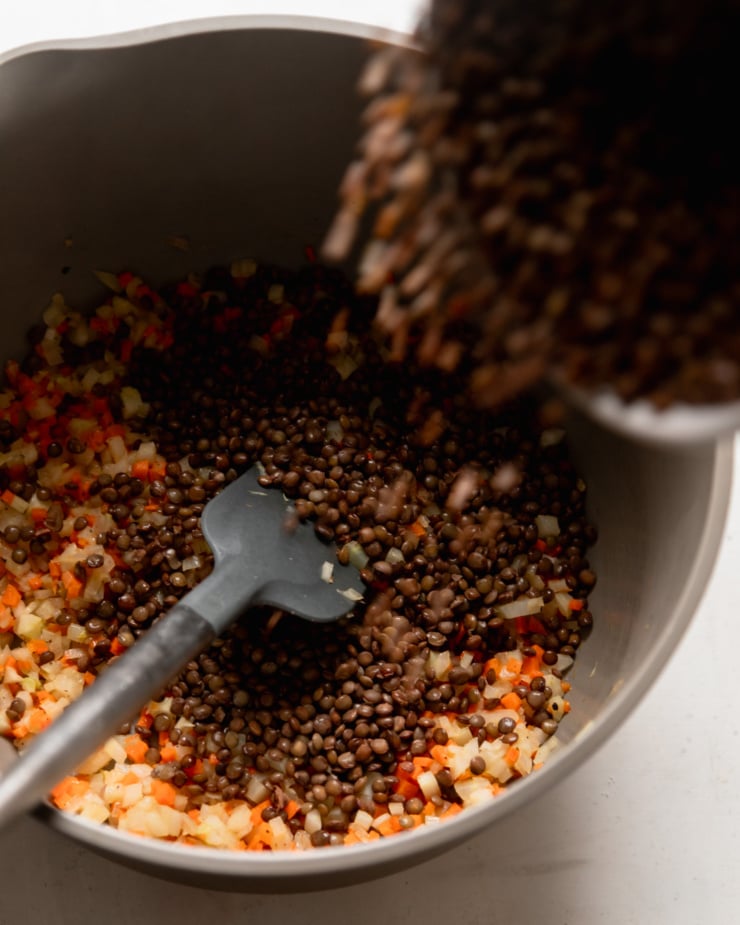 An overhead view shows a pot filled with sautéed fennel, carrots and green onions. Cooked French lentils are being poured into the pot.