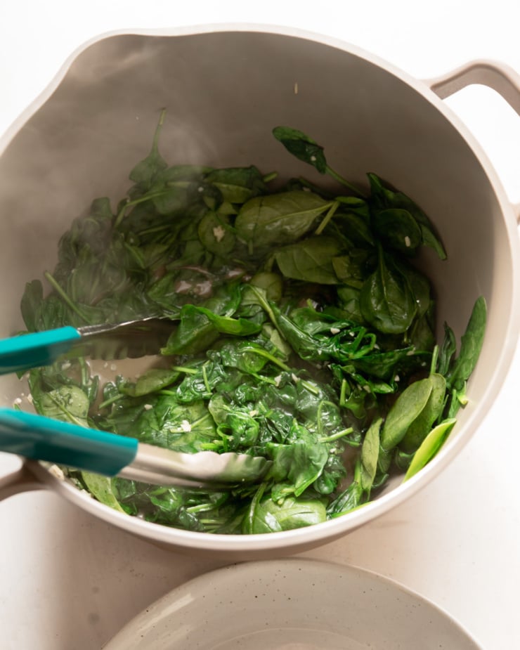 Top view shows wilted steamed bay spinach being placed in the pan using tongs.