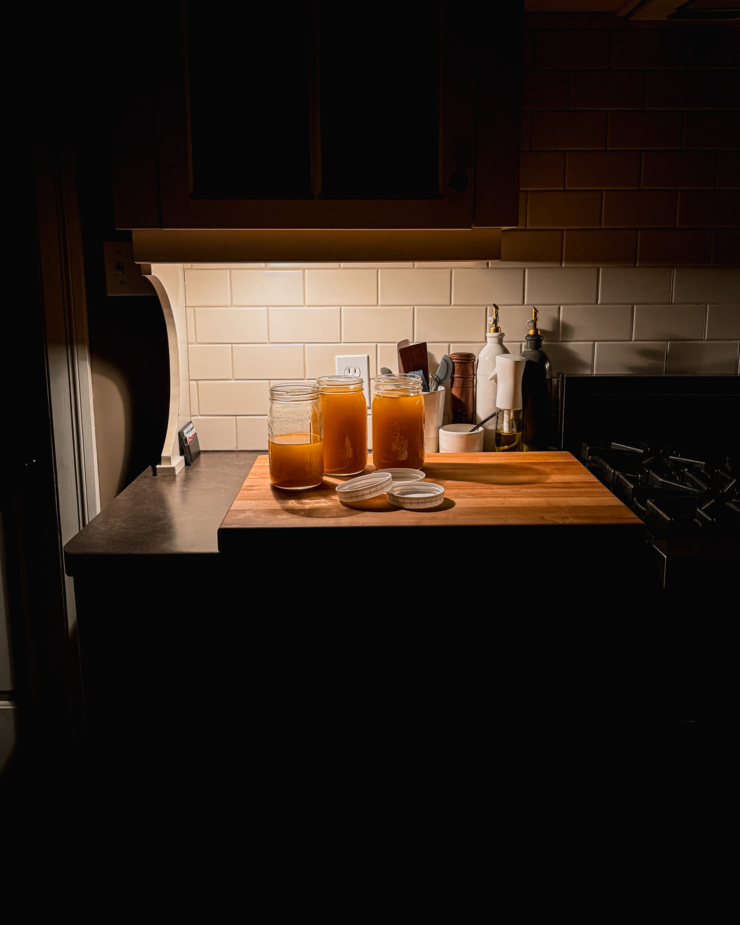 A head-on shot shows some jars of broth cooling on a kitchen counter, lit only by an under cabinet light in a dark kitchen.