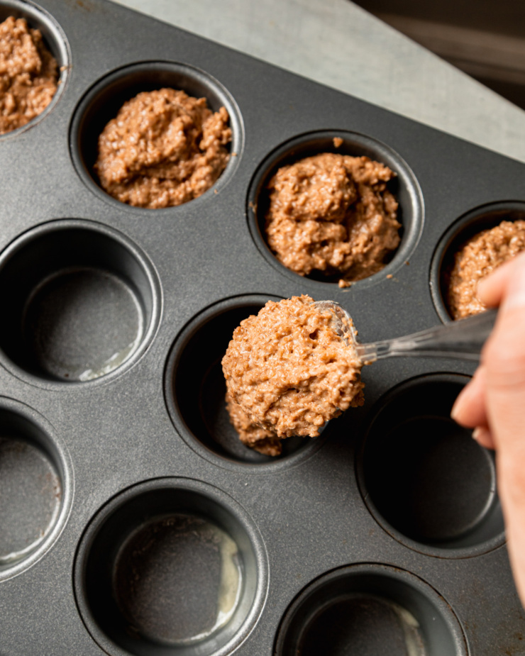An overhead shot shows a hand using a spoon to portion batter into a muffin tin.