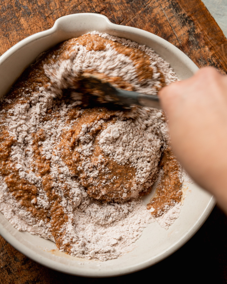 An overhead shot shows a hand using a spatula to bring the wet and dry components of a muffin batter together in a wide ceramic mixing bowl.