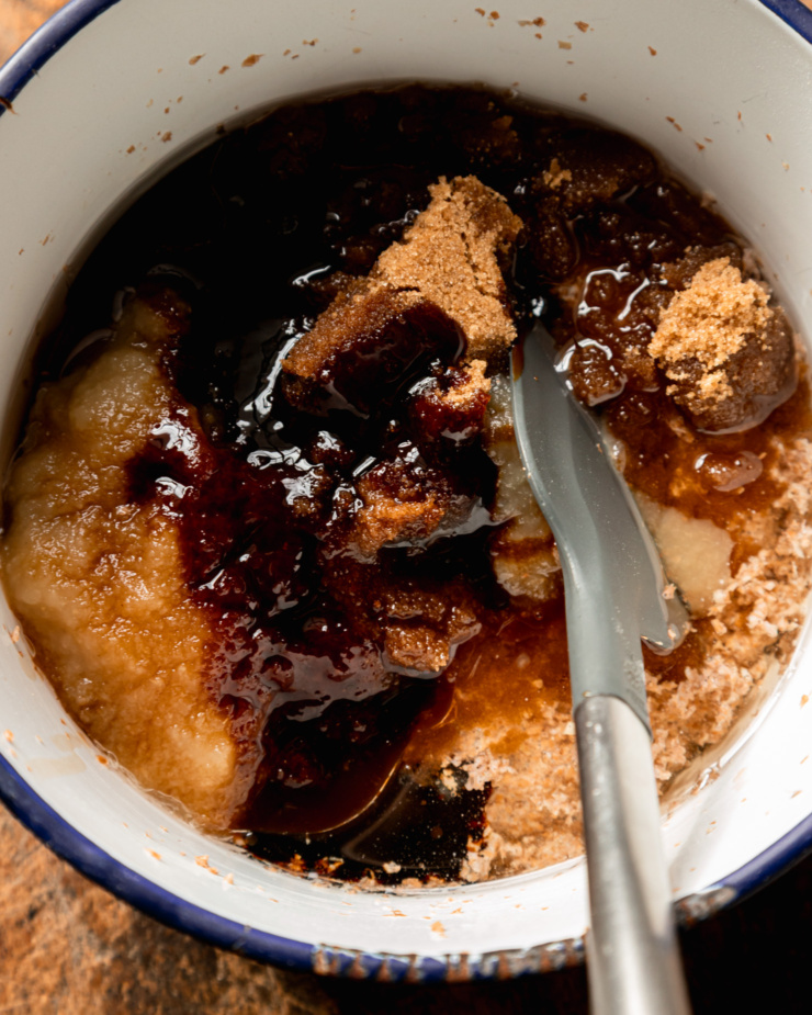 An overhead shot shows brown sugar, molasses, apple sauce, and oil on top of a soaked wheat bran mixture in a bowl. A spatula is sticking out of the mixture towards the camera.