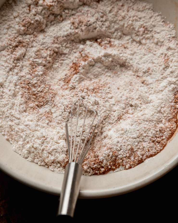 An overhead shot shows dry ingredients and a whisk in a wide mixing bowl.