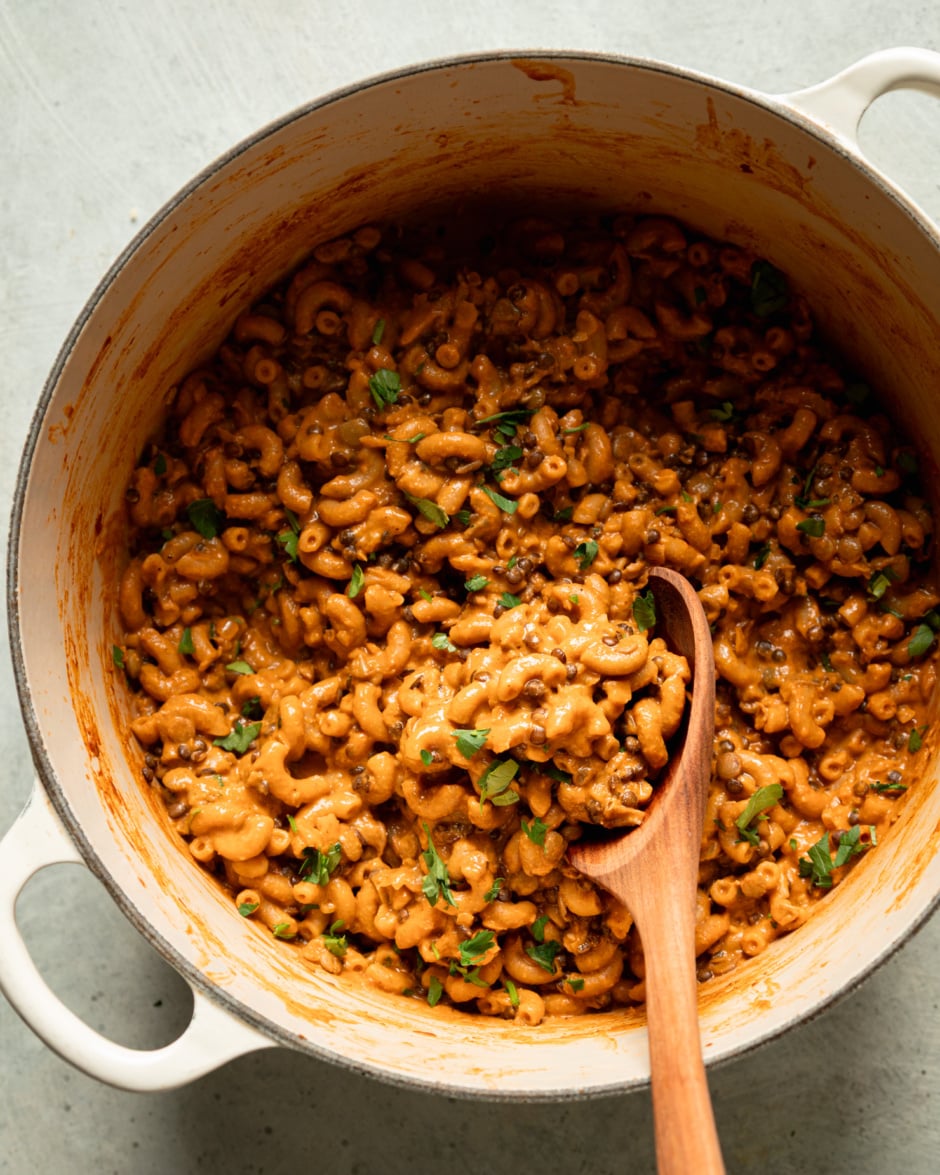 An overhead shot shows a Dutch oven filled with a vegan hamburger helper-style mixture with lentils, elbow macaroni, and chopped parsley. A wooden spoon is sticking out of the mixture.