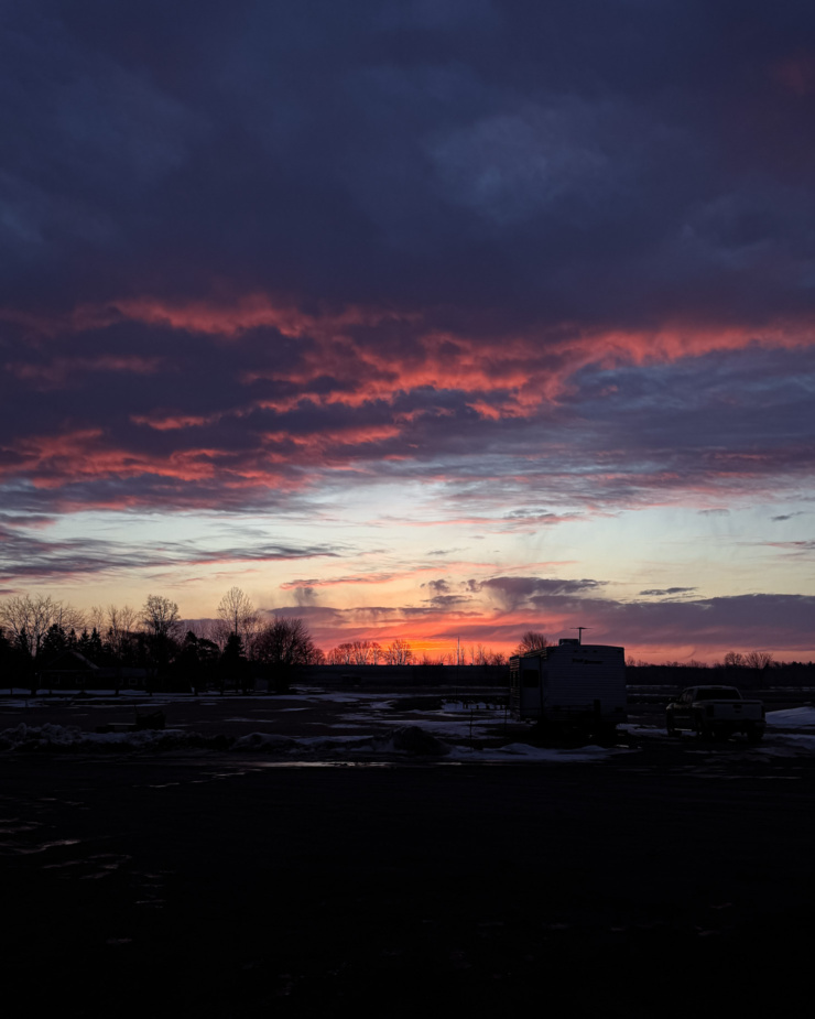 A head-on shot shows an early morning sunrise across a farmer's field. Some clouds are lit with bright salmon pink-hued light.