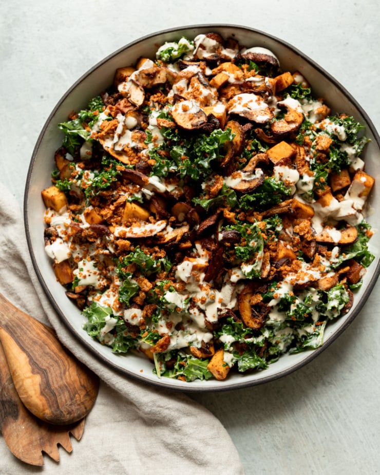An overhead shot shows a wide dish filled with roasted mushrooms and kale salad. The salad is topped with creamy garlic cashew dressing and seasoned bread crumbs. Salad tongs are seen to the side.