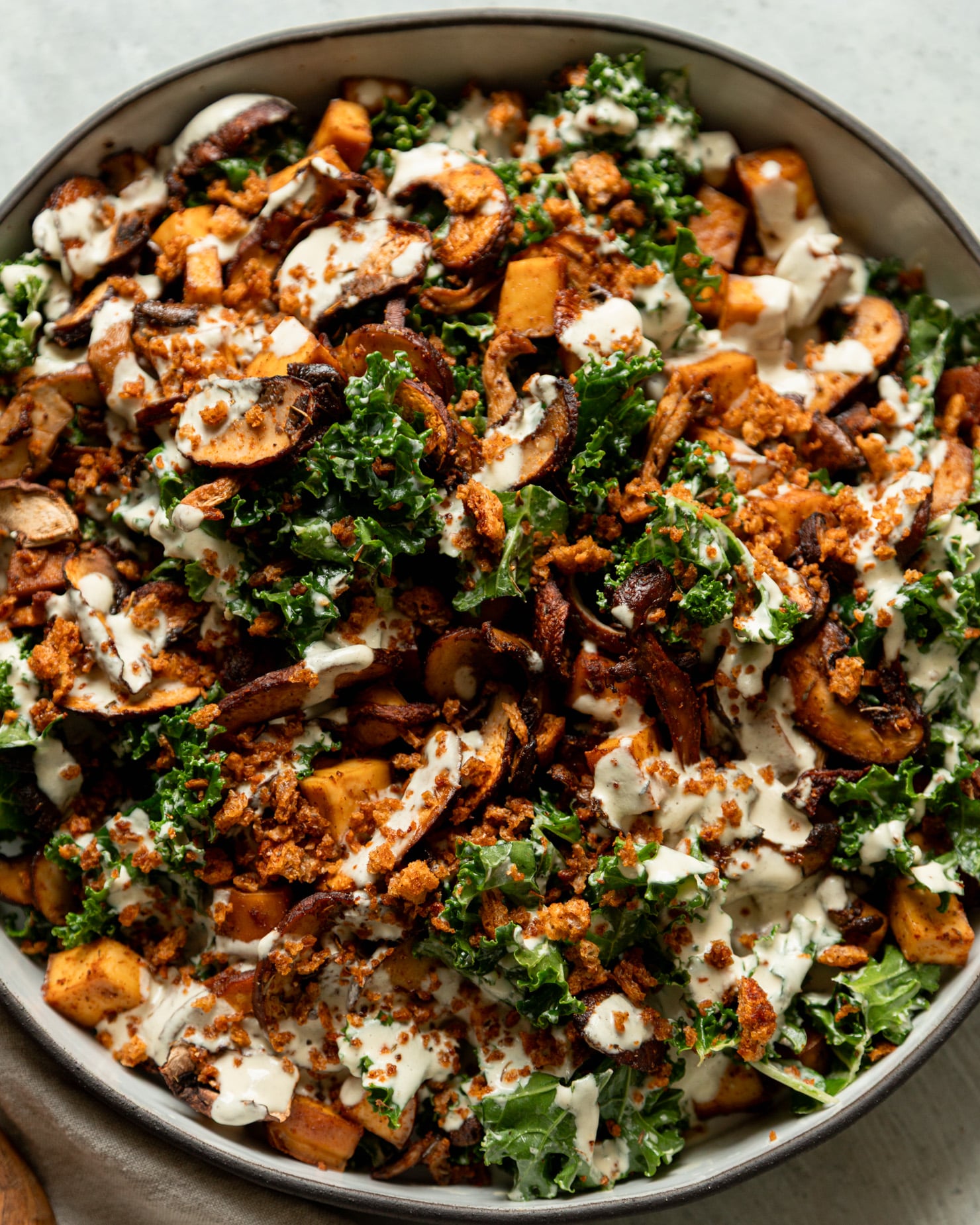 An up close, overhead shot shows a wide dish filled with roasted mushrooms and kale salad. The salad is topped with creamy garlic cashew dressing and seasoned bread crumbs