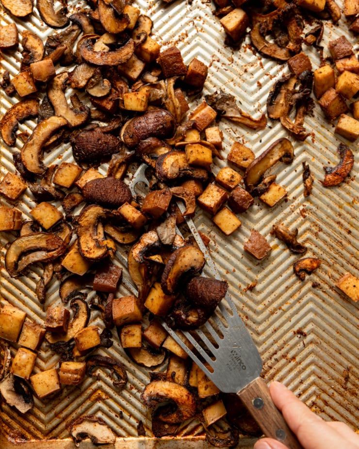 An overhead shot shows a hand using a fish spatula to lift up some roasted mushrooms and smoked tofu cubes from a baking sheet.