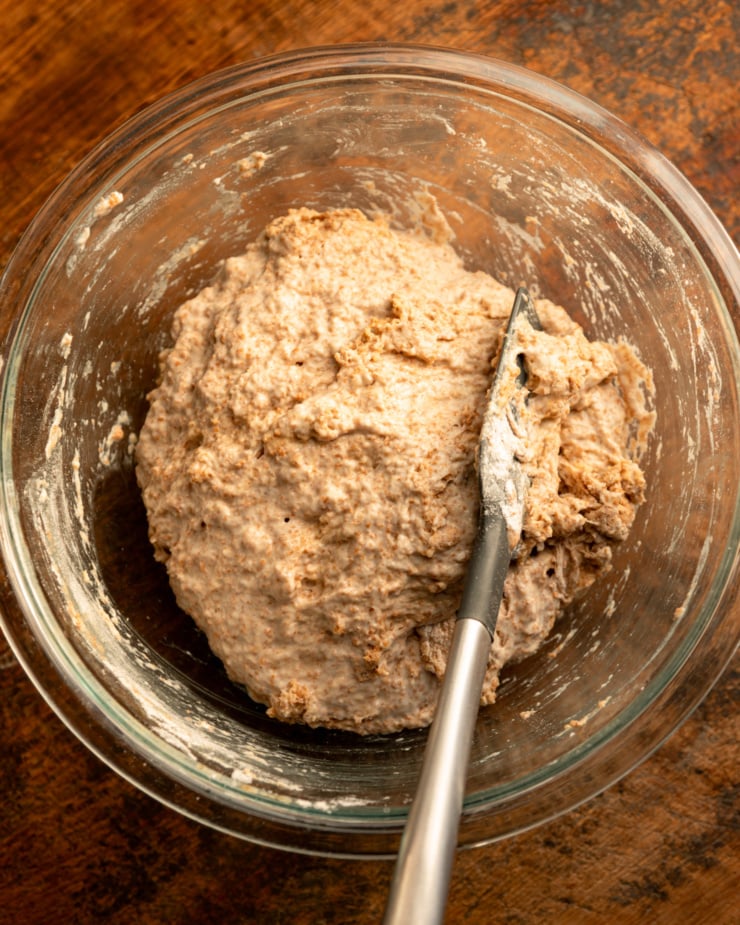 An overhead shot shows a clear glass mixing dough containing wheat bread dough and a spatula.