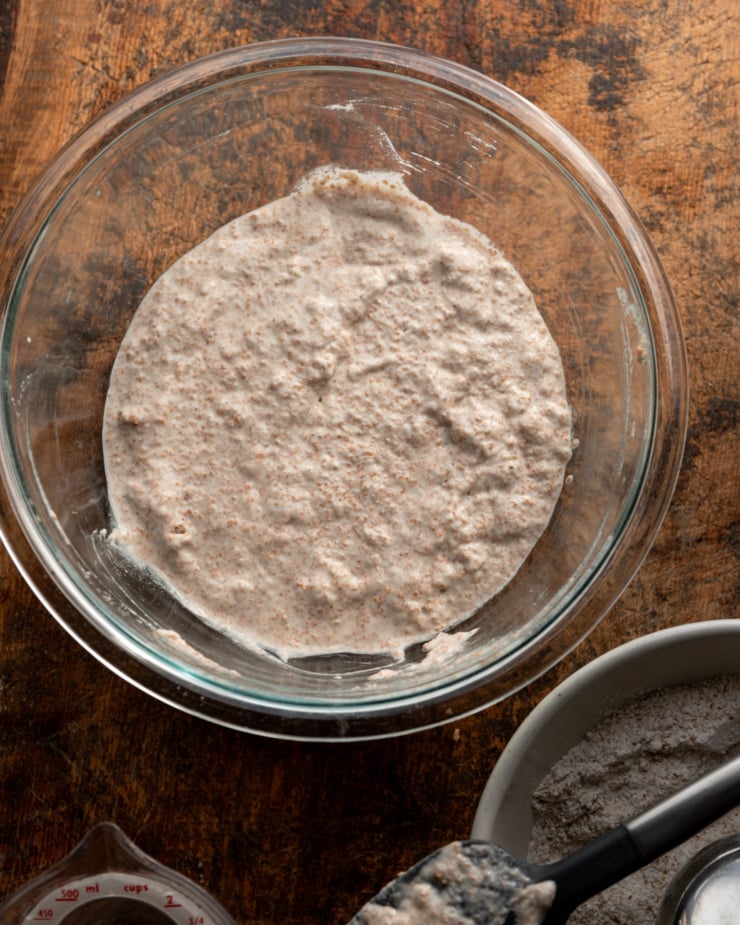 An overhead shot shows an autolyse mixture of whole wheat flour and water in a clear glass mixing bowl.
