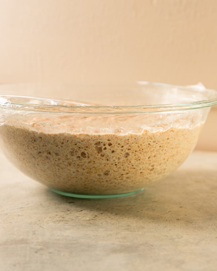 A head-on shot shows bread dough that has been left overnight in a clear bowl. There are lots of bubbles in the dough, seen through the clear glass.