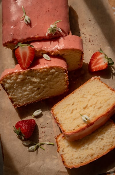 An overhead shot shows a sliced vegan lemon loaf with strawberry sumac glaze. Whole and halved strawberries as well as snow drop flowers are seen nearby. The photo is taken in warm sun light.