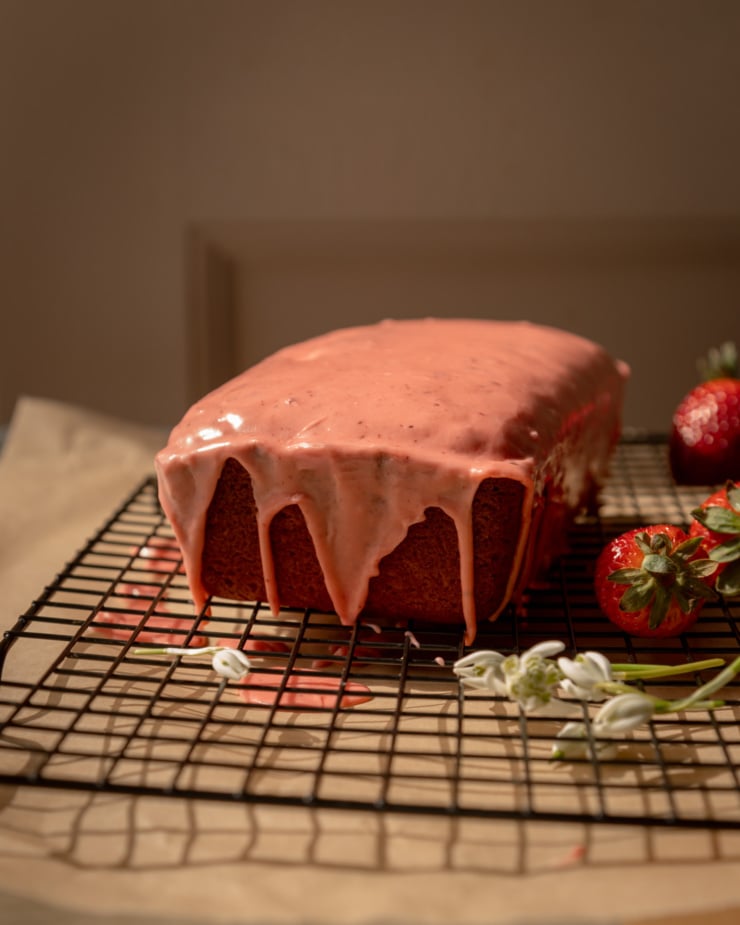 A head-on shot shows a vegan lemon loaf on a cooling rack with a strawberry sumac glaze spread over the top. Strawberries and snow drop flowers are nearby.