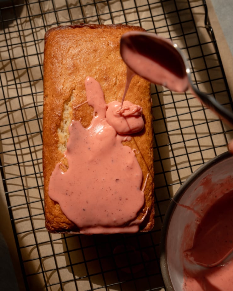An overhead shot shows a spoon drizzling strawberry sumac glaze over a vegan lemon loaf that is resting on a cooling rack.