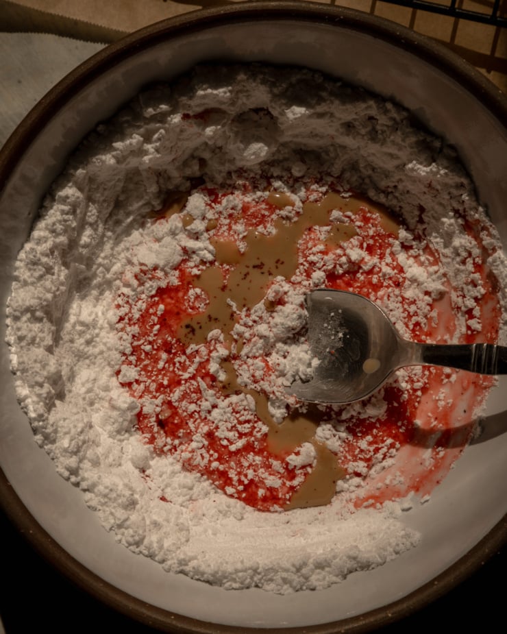 An overhead shot shows a bowl with powdered sugar, cooked strawberry juice, sumac, and tahini. A spoon sticks out of the mixture, ready to stir.