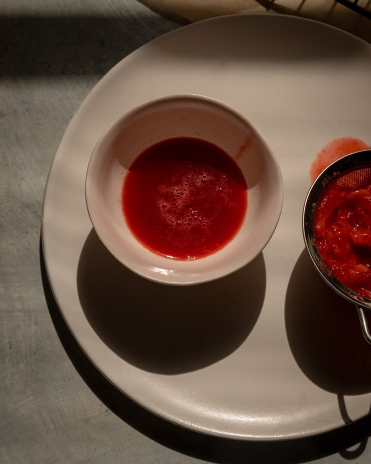 An overhead shot shows strained cooked strawberry juice next to a strainer with the strained fruit.