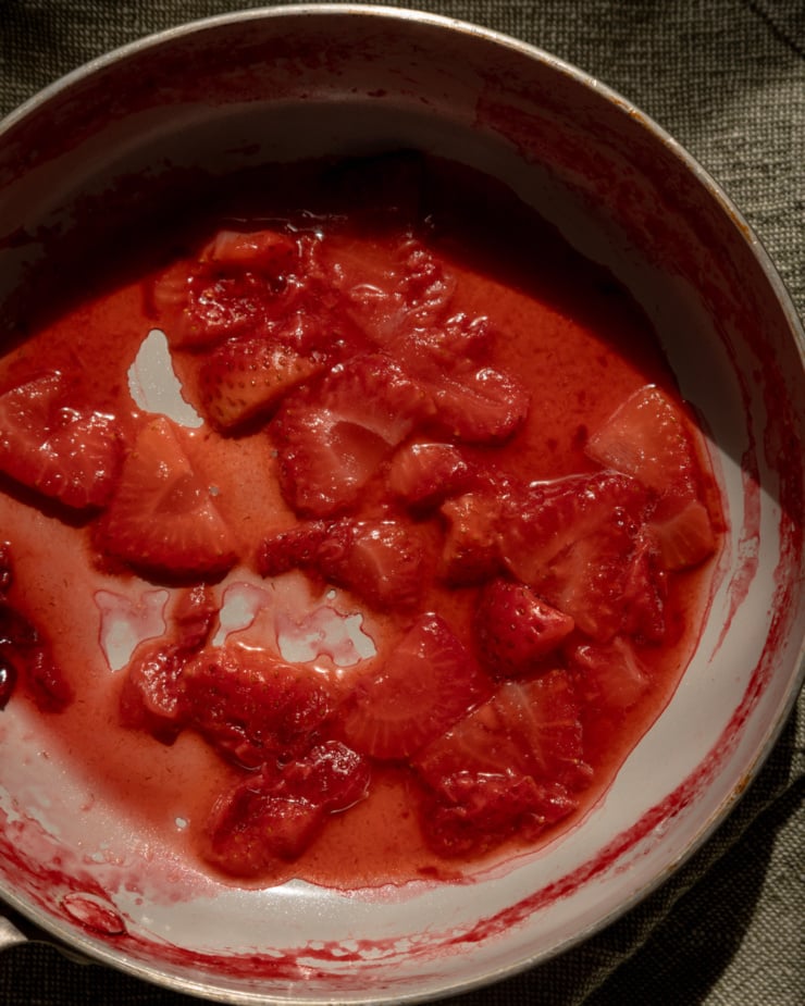 An overhead shot shows cooked sliced strawberries in a skillet with juices gathering around them.