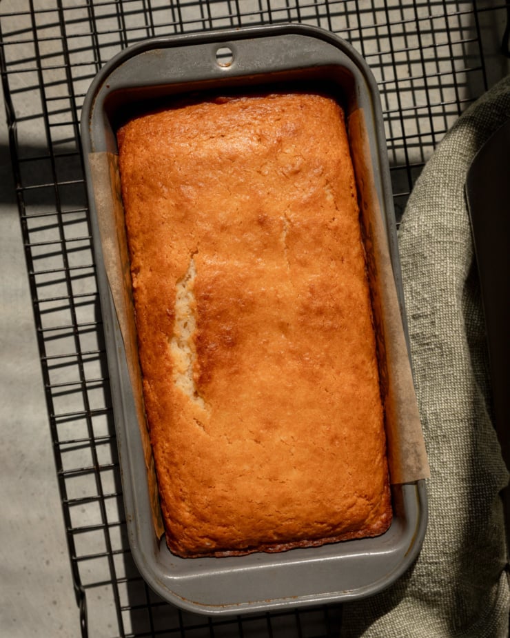 An overhead shot shows a freshly baked vegan lemon loaf in a loaf tin. It is set on a cooling rack in the sun.