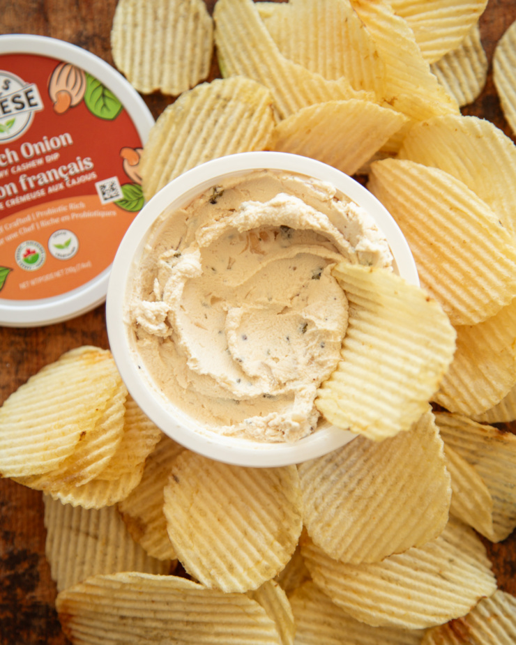 An overhead shot shows an open tub of Nuts for Cheese's French onion dip with rippled chips nearby. The dip is thick and creamy.