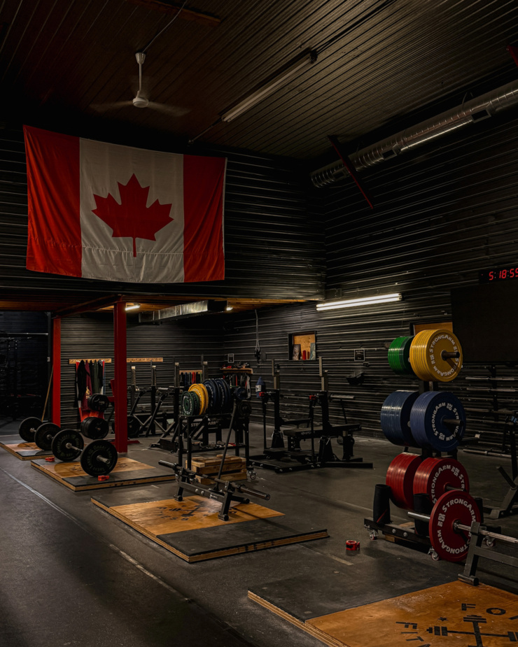 A head-on shot shows a room in a power lifting gym with a large Canadian flag draped on the wall.