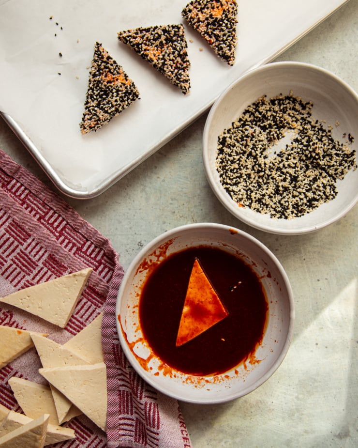 An overhead shot shows tofu being coated in a gochujang mixture, a bowl of sesame seeds, and a baking sheet with 3 sesame-coated pieces of tofu on it.