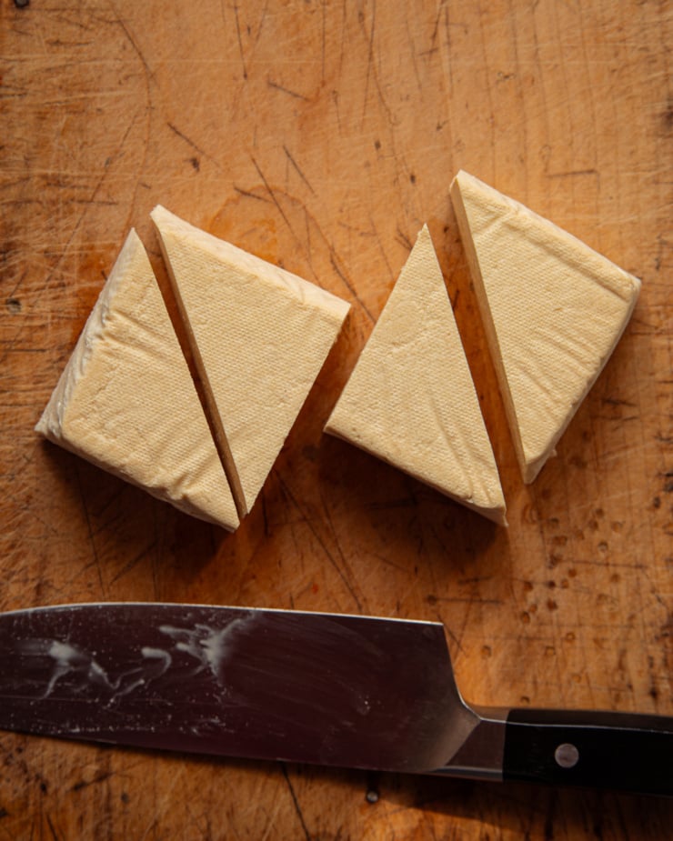 An overhead shot shows a tofu block cut into equal triangular quarters with a knife nearby.