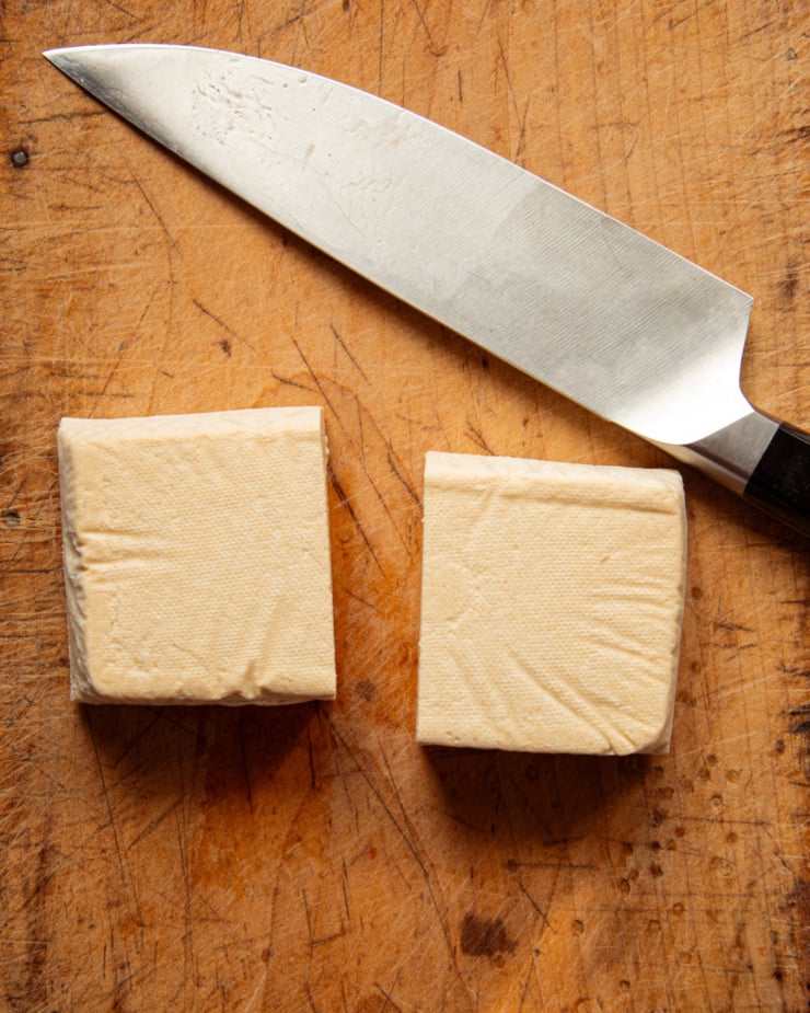 An overhead shot shows a block of extra firm tofu cut in half with a knife nearby.