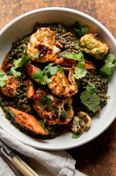 An overhead shot shows an individual serving of spinach and coconut stewed lentils with roasted carrots and cauliflower on top. The serving is garnished with fresh cilantro and chili crisp. A napkin and utensils are nearby.