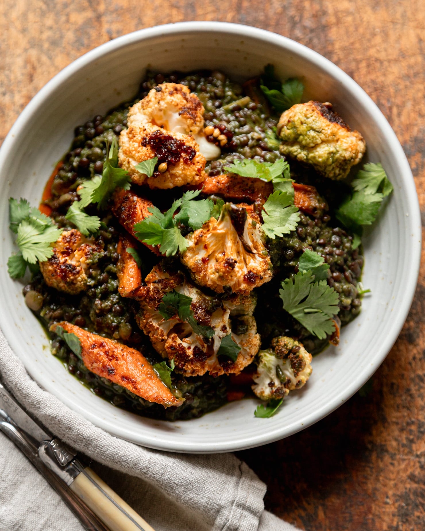 An overhead shot shows an individual serving of spinach and coconut stewed lentils with roasted carrots and cauliflower on top. The serving is garnished with fresh cilantro and chili crisp. A napkin and utensils are nearby.