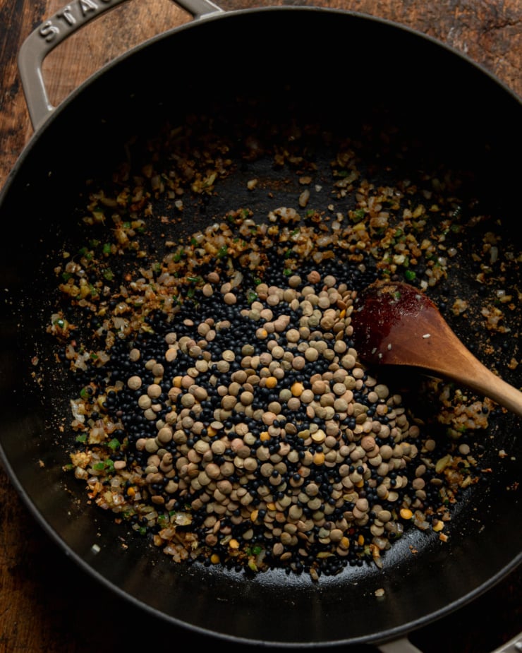 An overhead shot shows dry lentils added to a pot of sautéed shallots, ginger, garlic, jalapeño and spices.