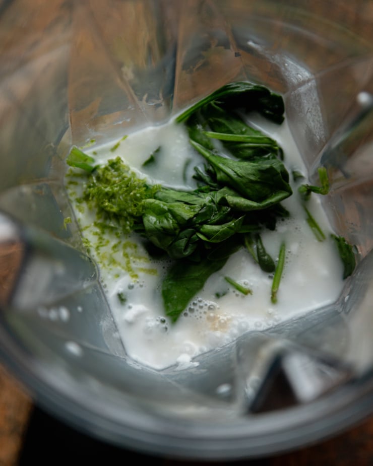 An overhead shot shows coconut milk in a blender with lime zest and wilted spinach.