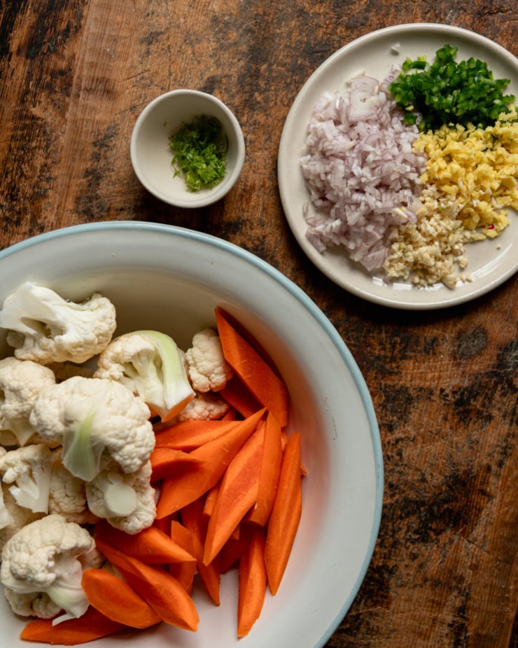 An overhead shot shows a plate with diced shallots, ginger, garlic, and jalapeño, a small bowl with lime zest in it, and a medium bowl with chopped carrots and cauliflower florets.