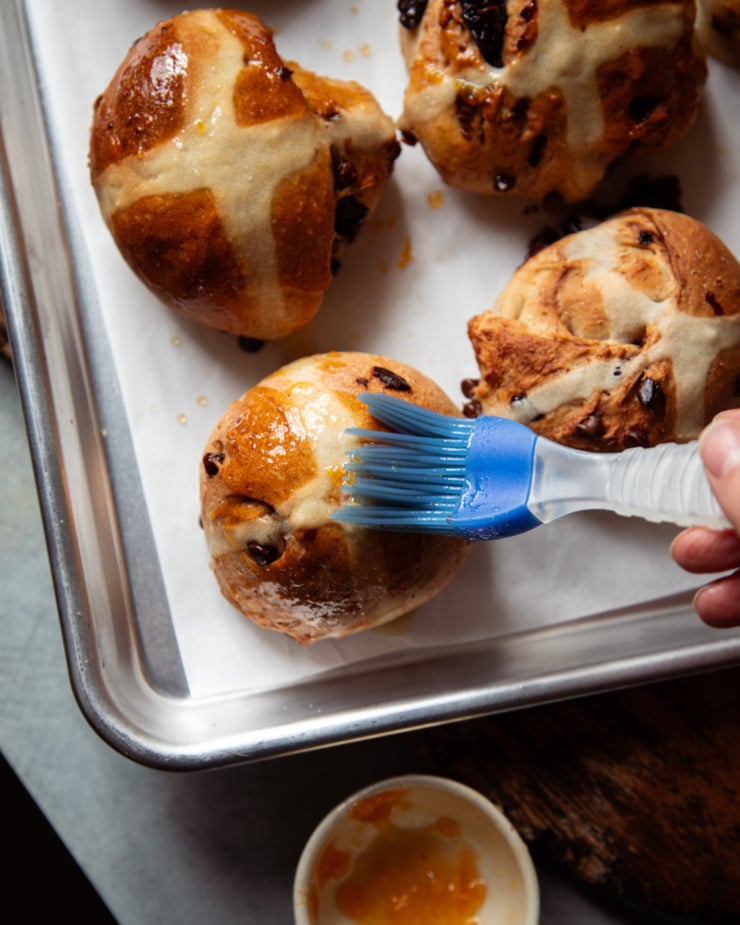 An overhead shot shows a hand using a brush to spread apricot glaze on some freshly baked vegan hot cross buns.