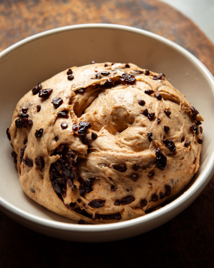 A 3/4 angle image shows vegan hot cross buns dough with dried cherries and chocolate chips after its first rise in a large ceramic bowl.
