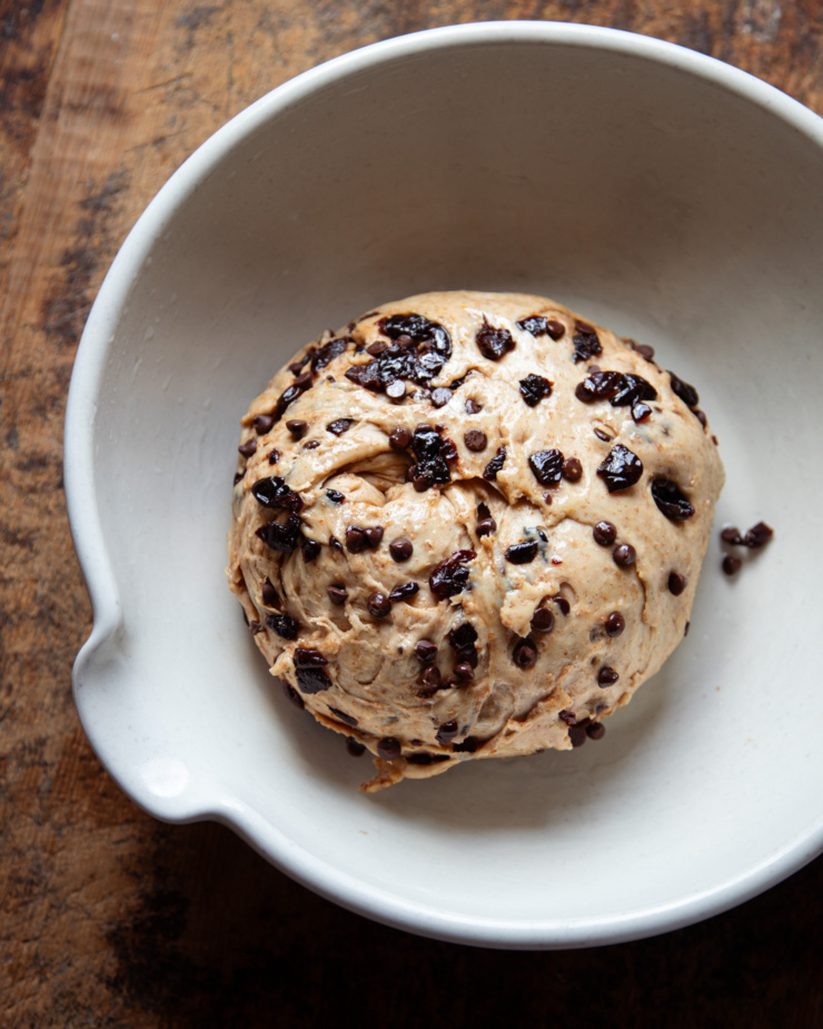 An overhead shot shows a soft dough in a large ceramic bowl. The dough is studded with chopped dried sour cherries and mini chocolate chips.
