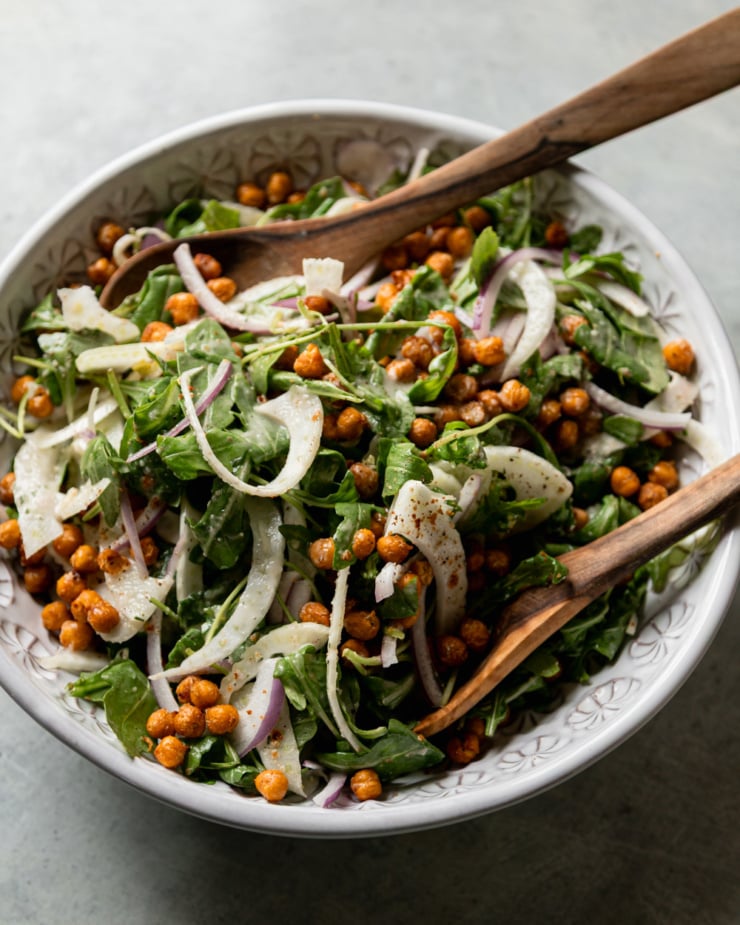 A 3/4 angle shot shows a chickpea arugula salad in a bowl with shaved fennel and red onions. There are wooden salad tongs sticking out of the salad.